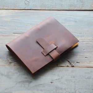 May include: A brown leather journal with a strap closure. The journal is lying on a wooden surface.