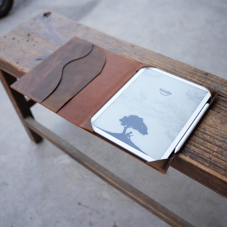 May include: A brown leather Kindle case with a matching flap. The Kindle is open, displaying a tree graphic and the Kindle logo. A white stylus rests on the side. The case is on a wooden surface.
