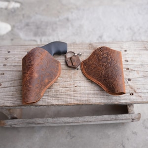 May include: Two brown leather holsters with floral tooling, one holding a black handgun. An antique padlock rests between the holsters on a weathered wooden surface. The holsters are designed to carry a firearm.