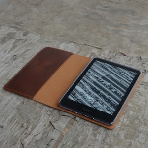 Puede incluir: Una funda de cuero marr&oacute;n con un interior beige est&aacute; abierta para revelar un lector electr&oacute;nico Kindle negro con una pantalla que muestra una imagen en blanco y negro.