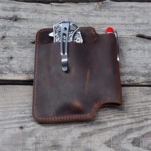 Könnte beinhalten: Ein brauner Leder-Taschenorganizer mit einem Fach für ein Klappmesser und einem Stifteinschub. Das Messer ist silberfarben mit einem dekorativen Muster und einem Clip. Der Stift ist rot.