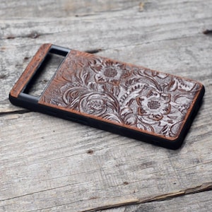 Puede incluir: Una funda de tel&eacute;fono de cuero marr&oacute;n con un dise&ntilde;o floral y un borde negro. La funda tiene un recorte para la lente de la c&aacute;mara.