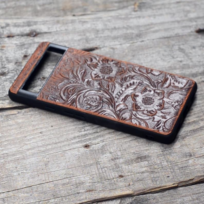 Puede incluir: Una funda de tel&eacute;fono de cuero marr&oacute;n con un dise&ntilde;o floral y un borde negro. La funda tiene un recorte para la lente de la c&aacute;mara.