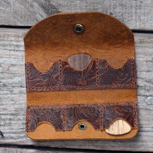Peut inclure: Un porte-médiators en cuir marron avec un motif en relief floral. Le porte-médiators a quatre fentes pour médiators et est ouvert pour révéler deux médiators en bois à l'intérieur.