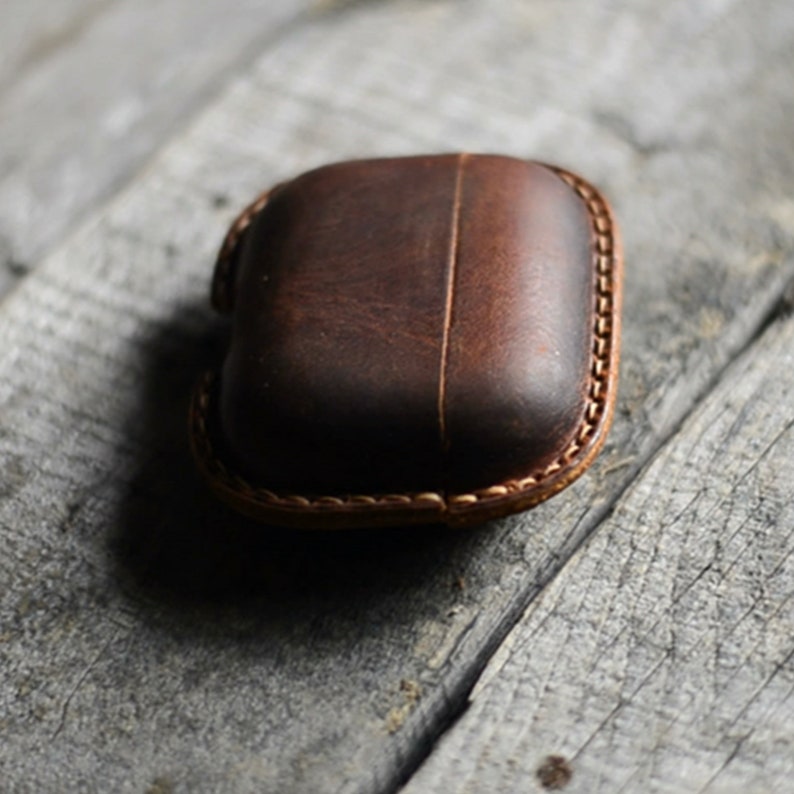 May include: A brown leather case, possibly for earbuds, rests on a weathered wooden surface. The case is square with rounded edges and a vertical seam down the center. The edges are stitched with a lighter brown thread.