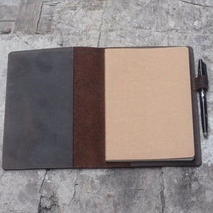 Puede incluir: Un cuaderno de cuero marrón con un portaplumas y una página en blanco abierta. El cuaderno tiene una cubierta marrón oscura y un interior marrón claro.