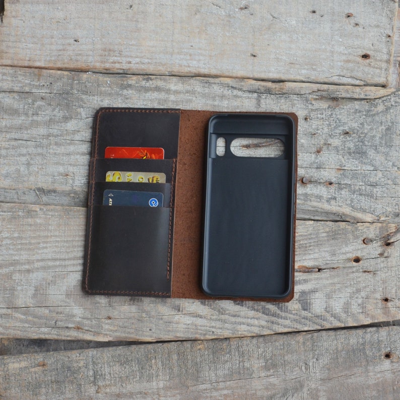 May include: A brown leather phone case with a flap that opens to reveal slots for credit cards. The case has a black phone holder and is closed.