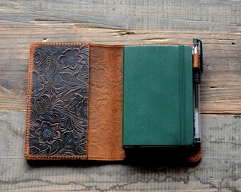 May include: A brown leather journal cover with a floral embossed design. The cover is open to reveal a green journal and a black pen.