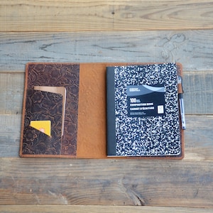 May include: A brown leather journal cover with a tooled floral design. The cover has a pocket on the left side and a pen holder on the right. A composition notebook is inside the cover.