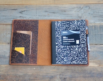 May include: A brown leather journal cover with a tooled floral design. The cover has a pocket on the left side and a pen holder on the right. A composition notebook is inside the cover.