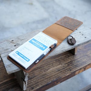 May include: A brown leather notebook cover with a "NEWS Professional REPORTER'S NOTEBOOK" inside. A black pen is secured in a leather loop. A small, rusty padlock rests nearby. The notebook is on a weathered wooden surface.