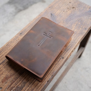 May include: A brown leather Bible cover with a debossed cross design. The cover is rectangular with a stitched border and a slightly distressed appearance. The Bible cover is resting on a wooden bench.