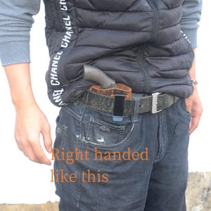 May include: A brown leather holster with a black clip is attached to a dark-coloured belt. A revolver is partially visible in the holster. The text "Right handed like this" is written in orange on the image. The person is wearing a black vest and blue jeans.