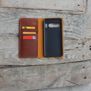 May include: A brown leather phone case with a flap that opens to reveal slots for credit cards. The case is closed and the phone is visible inside.