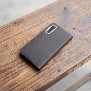 May include: A dark brown leather phone case with a rectangular camera cutout. The case is lying on a wooden surface with visible wood grain. The case appears to be closed and has a smooth, matte finish.
