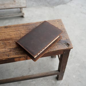 May include: A dark brown leather journal or notebook with a stitched border, resting on a rustic wooden bench. The journal's cover is a solid, rich brown, and the pages are visible along the edge. The bench has a weathered appearance.