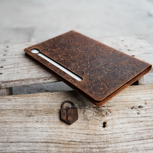 May include: A brown leather tablet case with a floral embossed pattern. The case features a stylus slot and sits on a wooden surface. A small, rusty metal padlock is also visible.