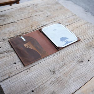 May include: A brown leather Kindle e-reader cover with a card slot. The cover is open, displaying the e-reader with a tree silhouette on the screen. The cover is on a weathered wooden table.
