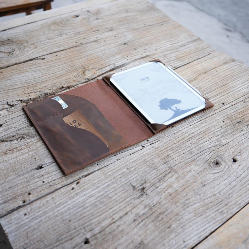May include: A brown leather Kindle e-reader cover with a card slot. The cover is open, displaying the e-reader with a tree silhouette on the screen. The cover is on a weathered wooden table.