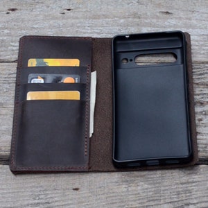 May include: A brown leather phone case with a built-in wallet. The case has slots for credit cards and a pocket for cash. The phone case is open, revealing a black phone inside.