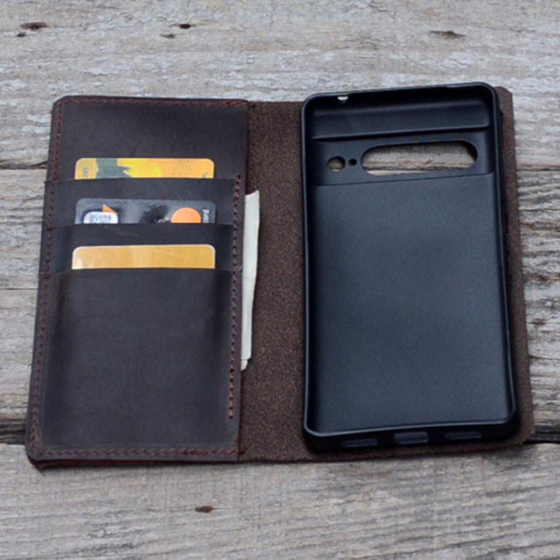 May include: A brown leather phone case with a built-in wallet. The case has slots for credit cards and a pocket for cash. The phone case is open, revealing a black phone inside.