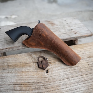 Op de afbeelding: Een zwart revolver is gedeeltelijk in een bruine leren holster gestoken met een bloemmotief. Een klein, antiek hangslot ligt op een houten oppervlak, naast de holster.