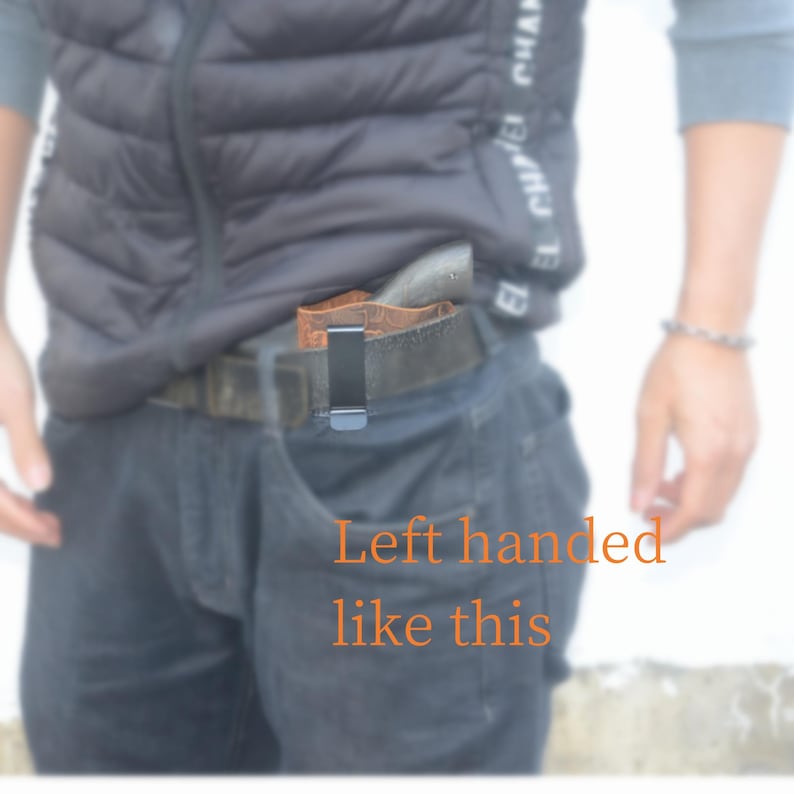 May include: A brown leather holster with a black metal clip is attached to a black belt. The holster is positioned inside the front pocket of dark blue jeans. The text "Left handed like this" is visible.