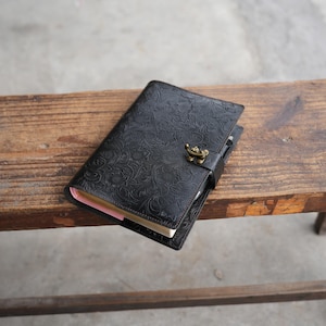 May include: Black leather journal with embossed floral pattern and a brass clasp. The journal is closed, with a pen tucked inside. The edges of the pages are visible, and the journal is resting on a wooden surface.