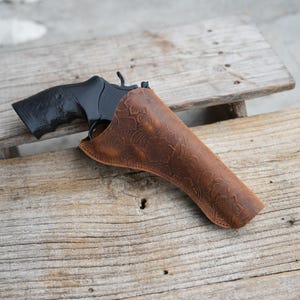 Puede incluir: Un revólver negro está parcialmente enfundado en una funda de cuero marrón. La funda presenta un diseño floral en relieve. La pistola y la funda descansan sobre una superficie de madera envejecida.