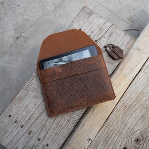May include: A brown leather e-reader case with a floral embossed design. The case is open, revealing the e-reader screen. A small, decorative metal piece is next to the case. The case is on a weathered wooden surface.