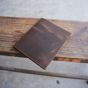 May include: A brown leather journal with a distressed finish, lying on a wooden bench. The journal has a rectangular shape with a stitched border. The leather has a mottled appearance with variations in color.
