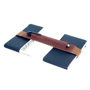 May include: A brown leather pen holder with a brown elastic band attached to a black notebook. The notebook has the text "LEUCHTTURM1917 DETAILS MAKE ALL THE DIFFERENCE" printed on the cover.