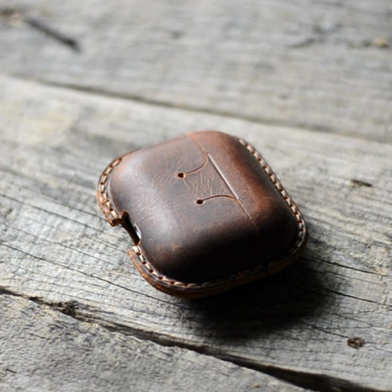 May include: A brown leather case for wireless earbuds. The case is rectangular with rounded edges and a stitched border. Two small holes and a decorative line are visible on the top. The case is sitting on a weathered wooden surface.