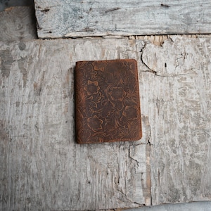 Puede incluir: Cartera de cuero marrón con diseño floral en relieve. La cartera rectangular está cerrada, con un patrón detallado de flores y hojas. El cuero tiene un aspecto envejecido, con variaciones de color y textura.