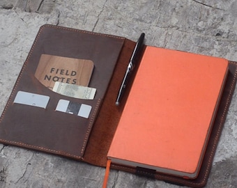 May include: A brown leather notebook cover with a pen holder, a pocket for a small notebook, and a pocket for cards. The cover is open to reveal an orange notebook.
