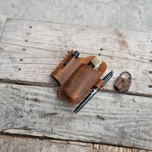 以下が含まれることがあります： 茶色の革製ペンホルダー。花柄のエンボス加工が施されており、ペン、小さなナイフ、懐中電灯が入っています。 小さな錆びた南京錠が、風化した木製の表面に置かれています。