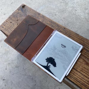 May include: A brown leather Kindle case with a pocket, open to reveal a Kindle device with a clear case and a stylus. The Kindle screen displays a black and white illustration of a tree and a person.