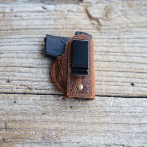 May include: A brown leather holster with a floral embossed design. The holster has a black metal clip and a brass-colored rivet. A black handgun is partially visible inside the holster. The holster is designed for concealed carry.