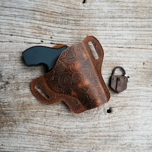 Könnte beinhalten: Ein braunes Lederholster mit floralem Prägemuster, das eine schwarze Handfeuerwaffe hält. Ein kleines, rostiges Vorhängeschloss liegt neben dem Holster auf einer verwitterten Holzoberfläche. Das Holster hat eine Gürtelschlaufe.