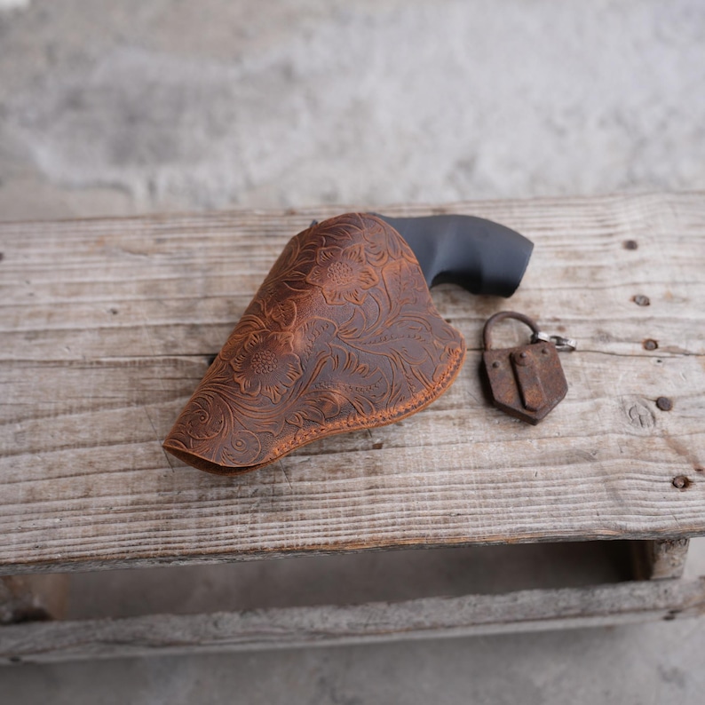 May include: A brown leather holster with a floral pattern, a black handgun, and a small, rusty padlock rest on a weathered wooden surface. The holster is intricately designed with embossed flowers and leaves.