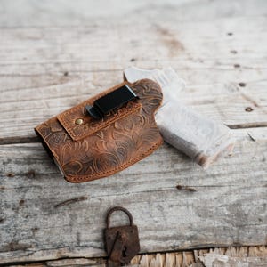 以下が含まれることがあります： フローラルエンボスデザインと黒い金属クリップが付いた茶色の革製ホルスター。ホルスターは木製の表面にあります。小さな錆びた南京錠も表示されています。