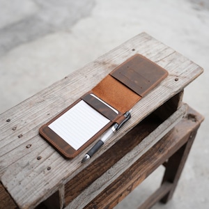 May include: Open brown leather notepad holder with a lined notepad and a pen. The notepad holder is open, revealing the interior with a card slot. The pen is silver and black. The notepad holder is on a rustic wooden surface.