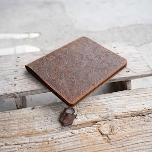 May include: A brown leather journal with a floral embossed design. The journal is rectangular and has a black border. A small, rusty padlock rests beside the journal on a wooden surface.