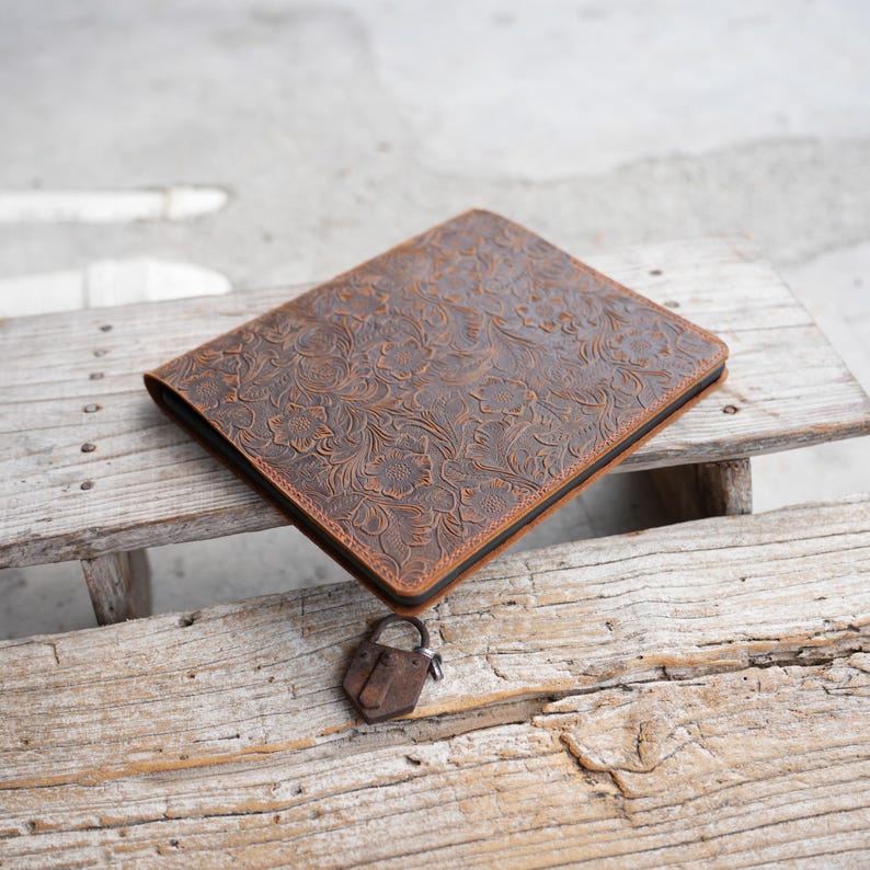 May include: A brown leather journal with a floral embossed design. The journal is rectangular and has a black border. A small, rusty padlock rests beside the journal on a wooden surface.