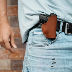May include: A brown leather holster holding a black handgun is placed in the front pocket of light blue jeans. The holster is secured by a black belt with a silver buckle. The person is wearing a light blue shirt.