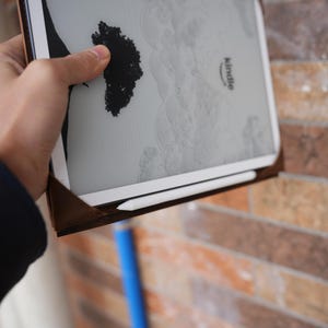May include: A Kindle e-reader with a white screen and a brown leather case. The screen displays a black and white image of a tree. A white stylus is placed along the bottom edge of the device. The word "Kindle" is printed on the device.