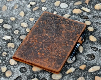 May include: A brown leather notebook with a floral embossed design. The notebook is lying on a gray surface with small stones.