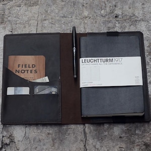 Op de afbeelding: Een bruine leren notitieboekhoes met een vak voor een pen, kaarten en een klein notitieboek. De hoes is open om een zwart notitieboek te laten zien met de tekst "LEUCHTTURM1917" en "DETAILS MAKE ALL THE DIFFERENCE." op de voorkant.