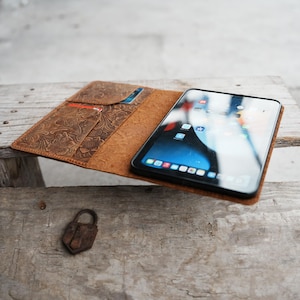 Puede incluir: Una funda de tableta de cuero marrón con un diseño floral en relieve, abierta para revelar una tableta con un bisel negro. La funda tiene ranuras para tarjetas y está sobre una superficie de madera. Un pequeño candado oxidado está al lado de la funda.