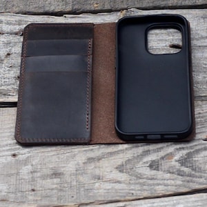 May include: A brown leather phone case with a card holder. The case is open, revealing a black phone case inside.